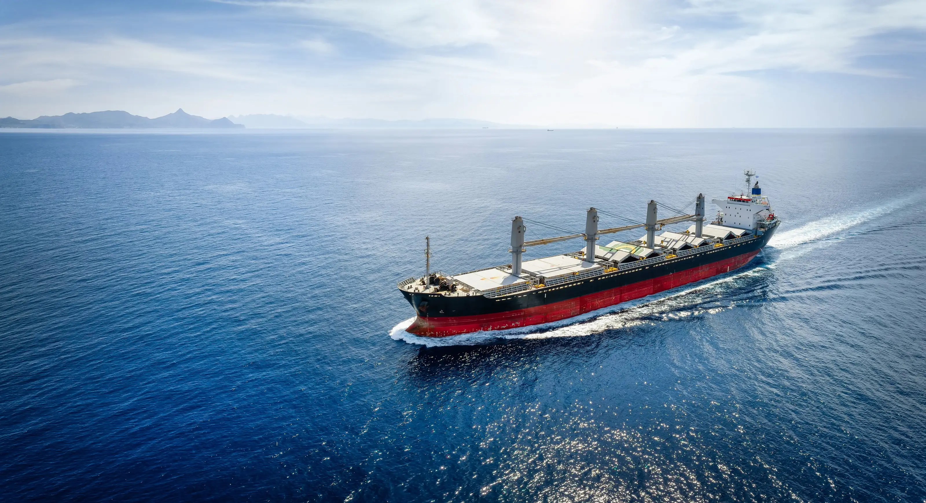 Aerial view of a loaded bulk carrier under way in open sea, deck cranes amidships and a white wake astern.