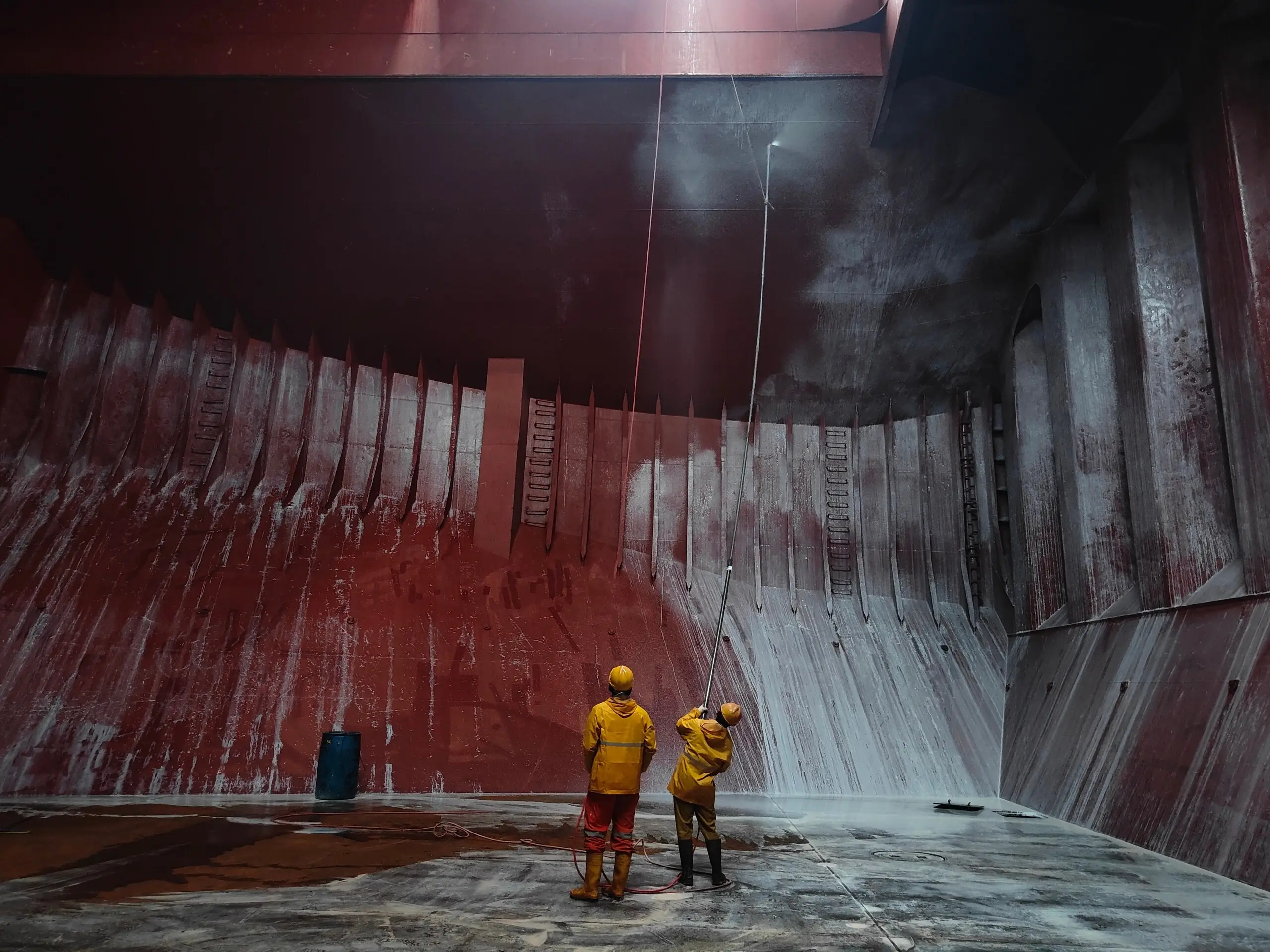 Two technicians in yellow protective gear pressure-washing the bulkhead of a bulk carrier hold, half of it already washed down to bare steel.