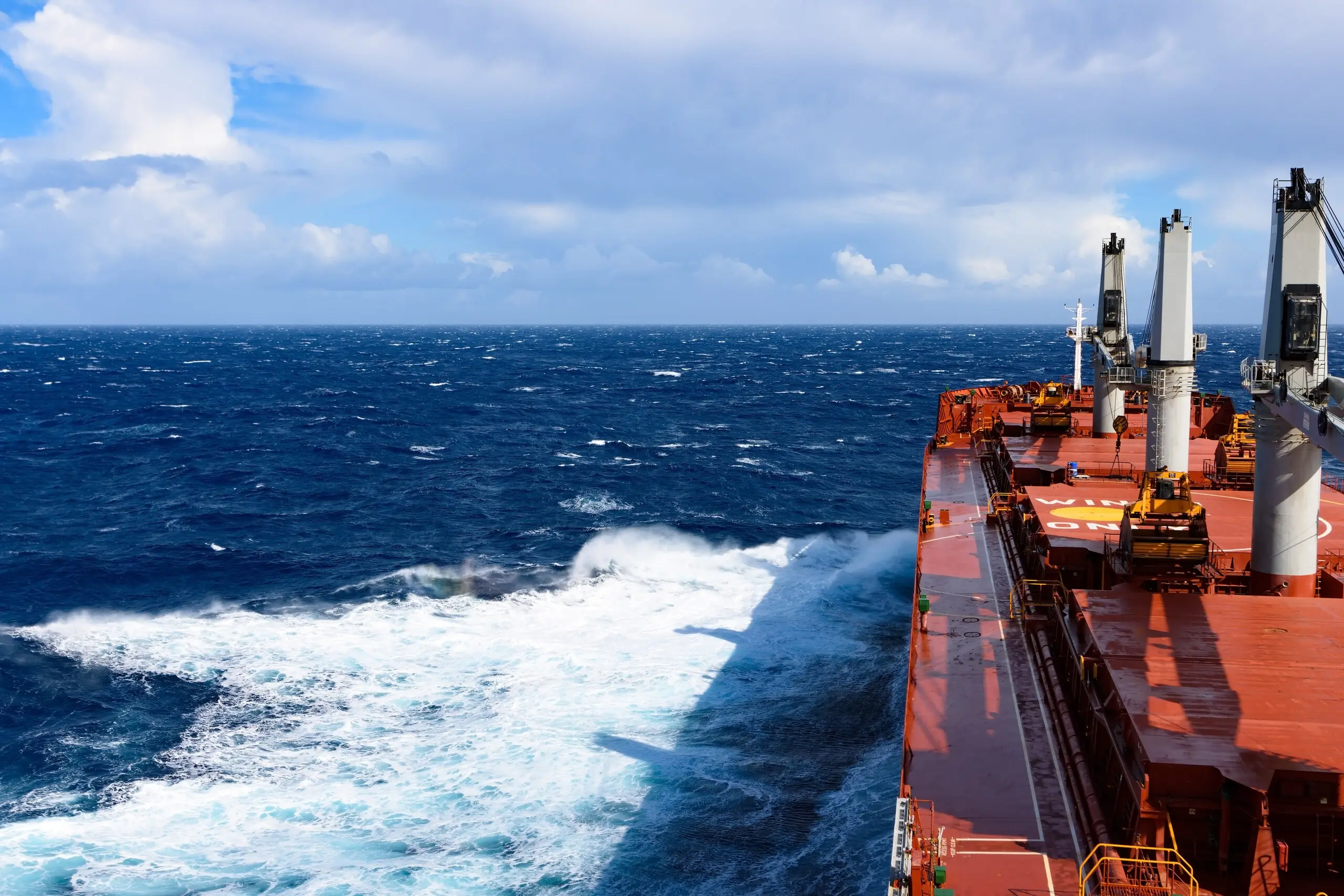 View forward from a bulk carrier's superstructure — deck cranes and hatch covers to starboard, open ocean to port.